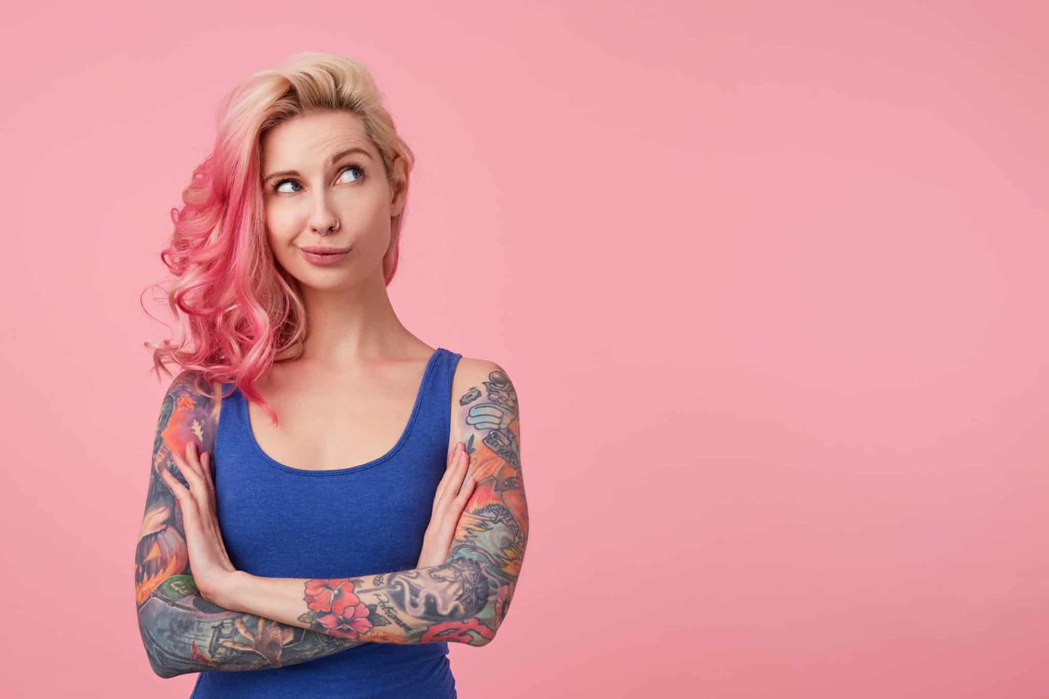 Woman with blond and pink hair and tattoed arms looking up to the right corner thinking of something.